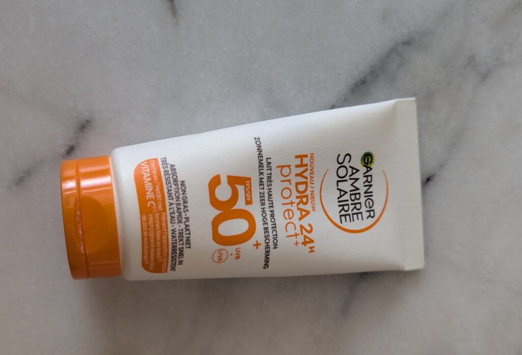 Photographie d'un tube de crème solaire Garnier Ambre Solaire Hydra 24H Protect+, SPF 50+, à la vitamine C, avec bouchon orange.