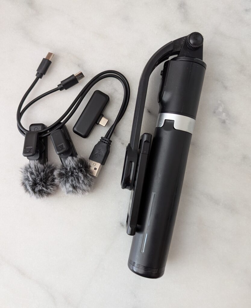 Photographie d'une perche à selfie/trépied pliable noire et pliée, accompagnée d'un support pour smartphone, ainsi que de deux micros cravate avec bonnettes anti-vent en fourrure grise, un adaptateur USB-C et des câbles de raccordement.