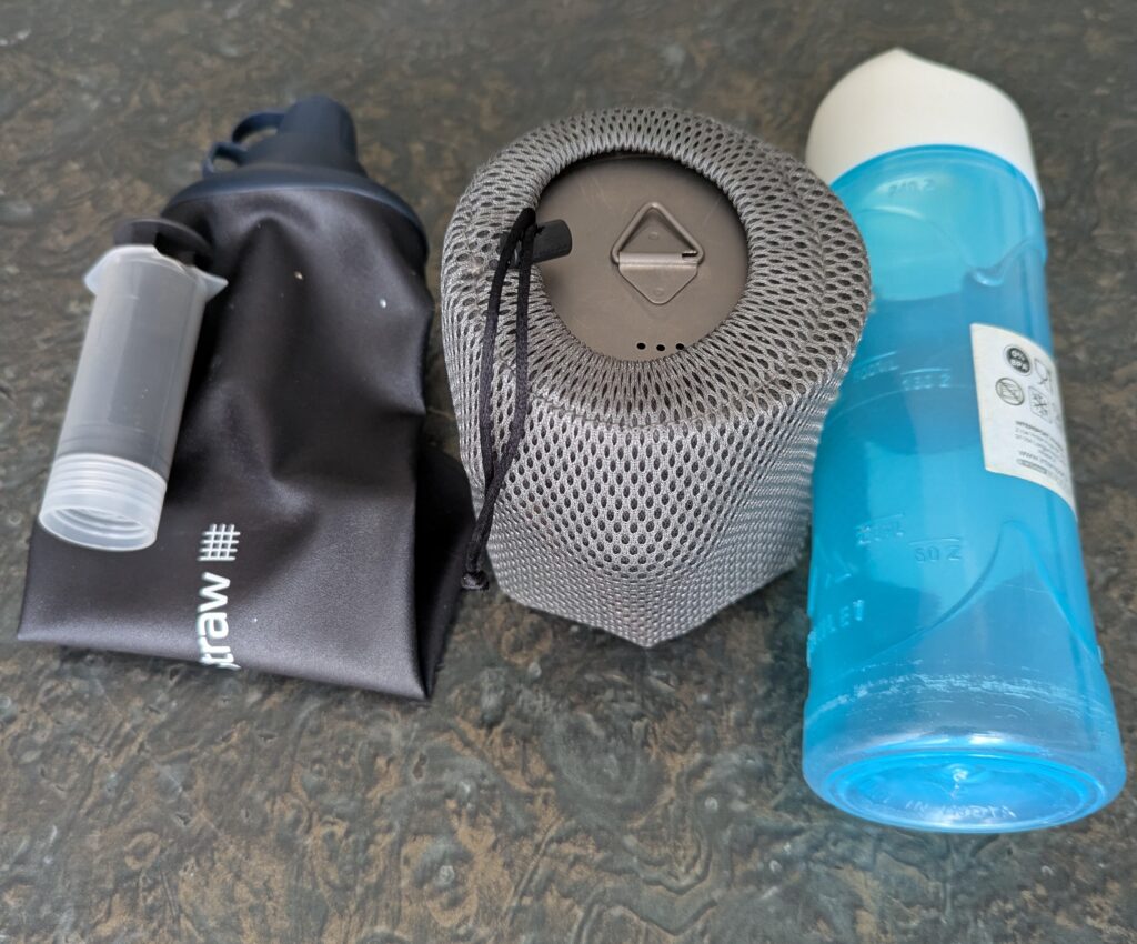 Photographie de trois éléments posés côte à côte : à gauche, une gourde filtrante souple noire LifeStraw avec sa seringue de rinçage ; au centre, une housse en filet gris contenant la gamelle titanium avec l'ensemble du petit matériel de cuisine (réchaud, cartouche de gaz, couverts, couteau, briquet, allumettes) ; à droite, une gourde rigide bleue McKinley 0,75L.