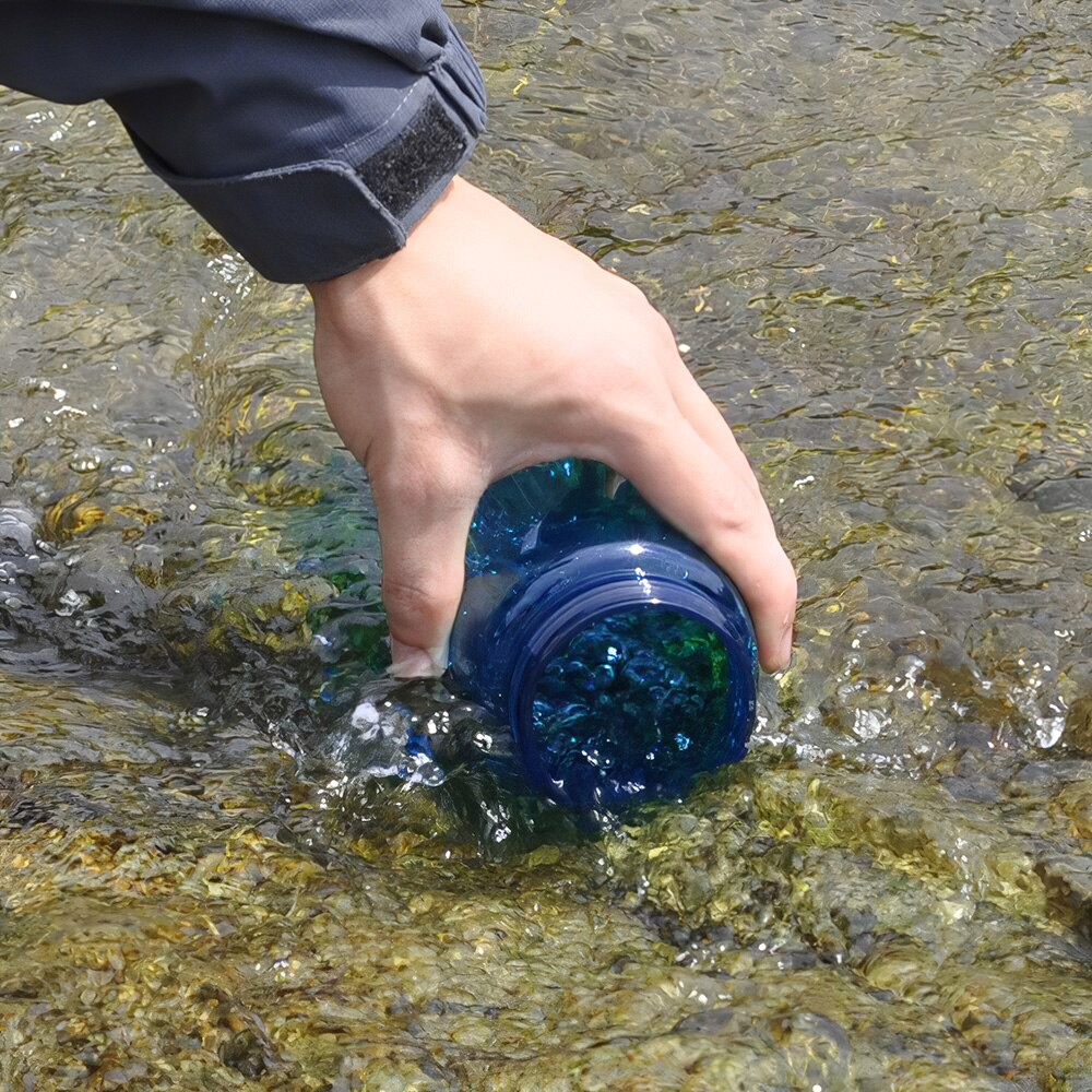 Une main remplit une gourde bleue transparente en la plongeant dans l’eau claire d’un cours d’eau peu profond, sur un lit de galets.