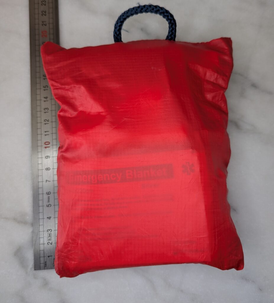 Photographie de la trousse de secours rangée, une pochette rouge en tissu et fermée, posée à côté d'une règle graduée indiquant une hauteur d'environ 23 cm.