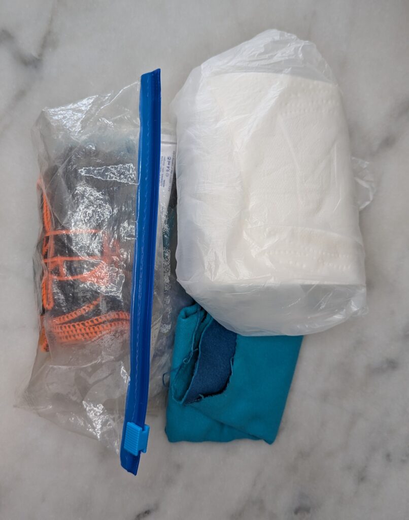 Photographie de deux sachets zippés transparents posés côte à côte : l'un contenant tous les accessoires de toilette, l'autre contenant un rouleau de papier toilette compressé et un chiffon microfibre bleu à part.