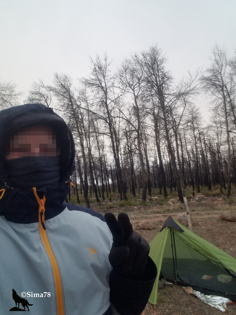 Randonneur emmitrouflé dans une veste et un cache-cou, faisant le signe de la victoire, devant une tente légère verte plantée au milieu d'une forêt de troncs calcinés.
