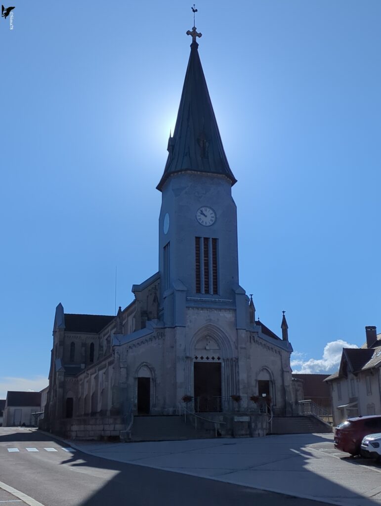 Église en pierre à façade néogothique, dominée par un clocher à flèche pointue couverte de métal vert-de-gris, surmontée d’une croix et d’un coq, avec une horloge sous la flèche et un escalier d’accès précédé d’une place, en contre-jour sous un ciel bleu.
