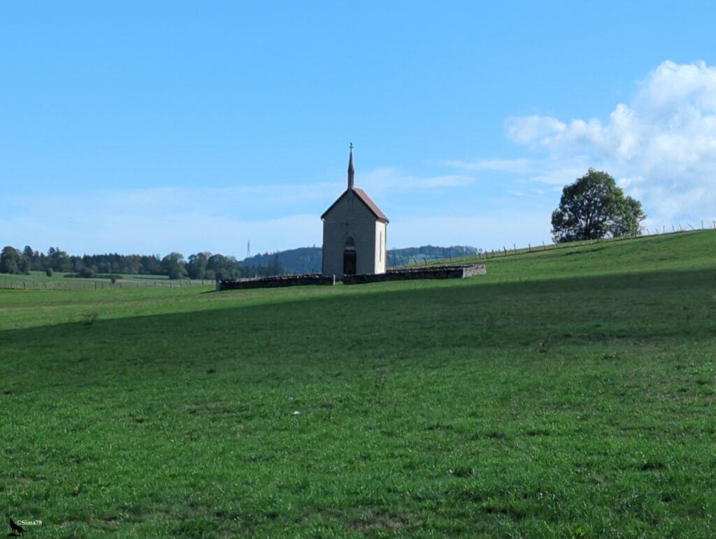 Petite chapelle de pierre claire à toit de tuiles rouges et clocheton surmonté d’une croix, entourée d’un muret bas, isolée au sommet d’une prairie verte, avec un arbre à droite et des collines boisées au loin sous un ciel bleu.