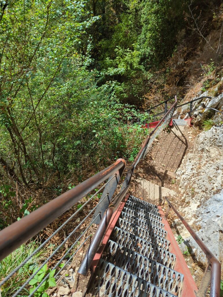 Vue plongeante sur un escalier métallique à marches ajourées et à structure rouge, équipé de rampes et de câbles tendus, qui descend le long d’une paroi rocheuse claire, au milieu d’une végétation dense.
