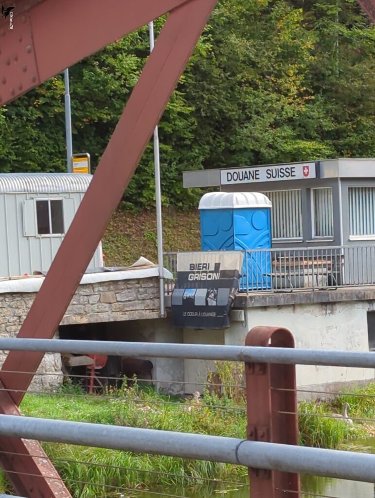 Vue à travers la structure métallique rouge d’un pont sur un poste de douane suisse, avec l’enseigne « Douane suisse » et un drapeau à croix blanche, une cabine sanitaire bleue, une banderole publicitaire et une roulotte de chantier, au pied d’un talus boisé.