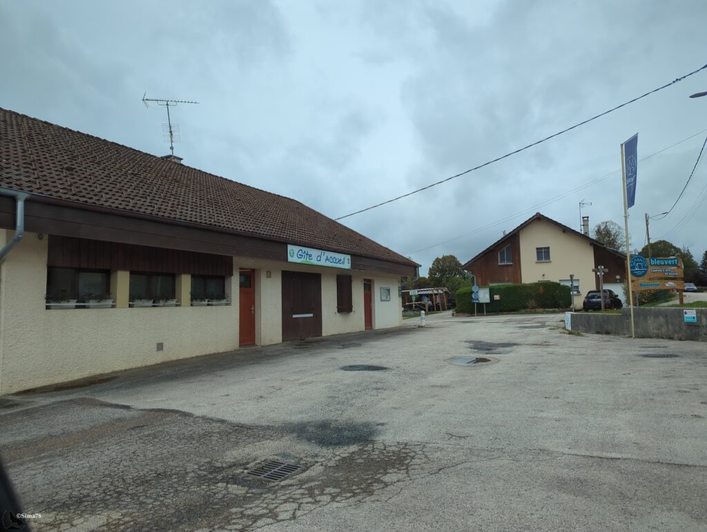 Bâtiment bas aux murs crépis beige et à la toiture de tuiles brunes, portant l’enseigne « Gîte d’accueil », devant un parking en enrobé humide, avec un panneau « Camping & Gîte de Maîche » à droite sous un ciel nuageux.