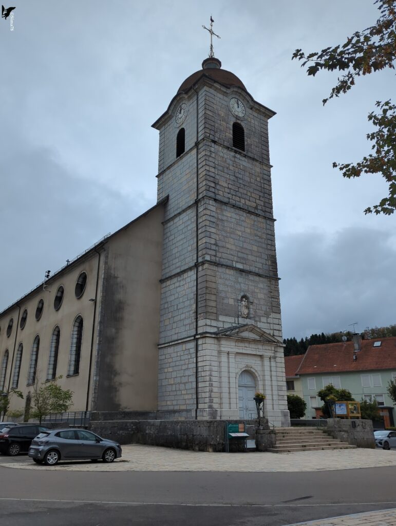 Église en pierre dont le haut clocher carré, coiffé d’un dôme de tuiles et d’une croix surmontée d’un coq, domine une nef à fenêtres arrondies, avec un escalier d’accès et des voitures garées au premier plan, sous un ciel gris.