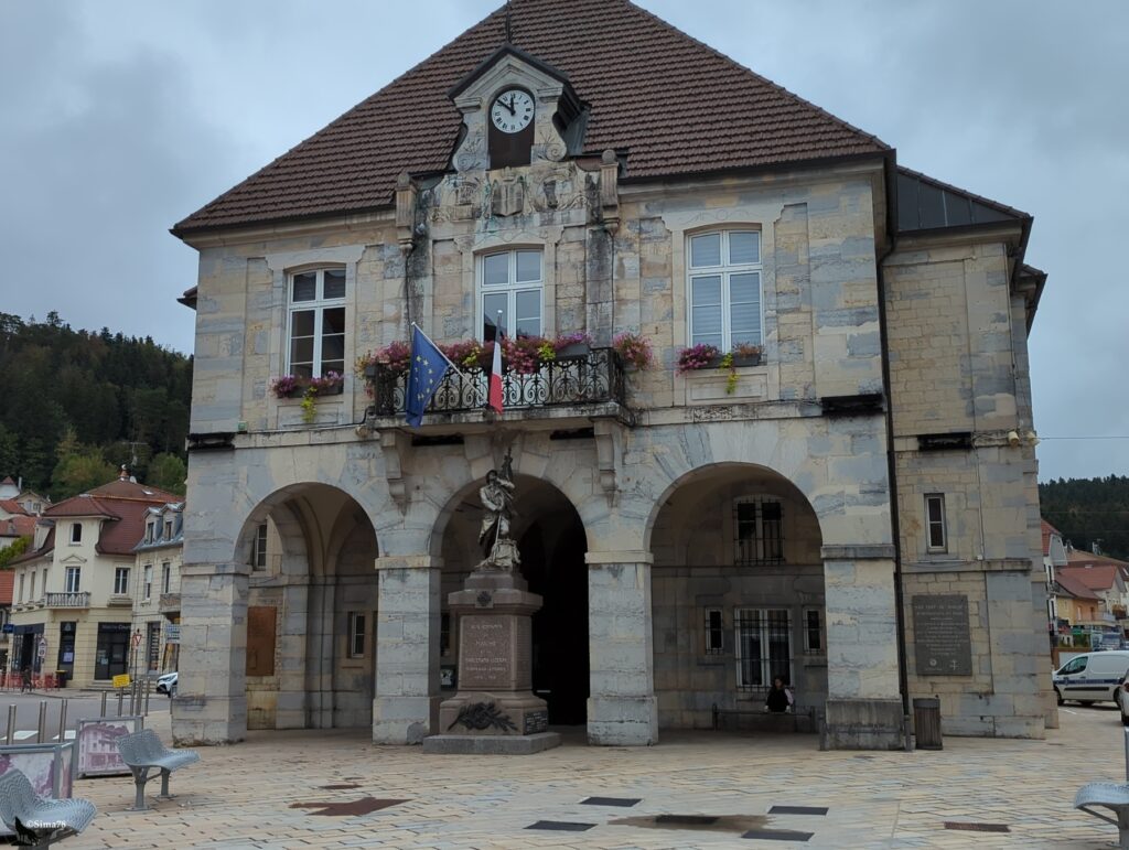 Façade en pierre claire d’une mairie à toit de tuiles brunes et horloge en pignon, avec un balcon en fer forgé fleuri portant les drapeaux européen et français, et un monument aux morts sous l’arcade centrale, sur une place pavée.