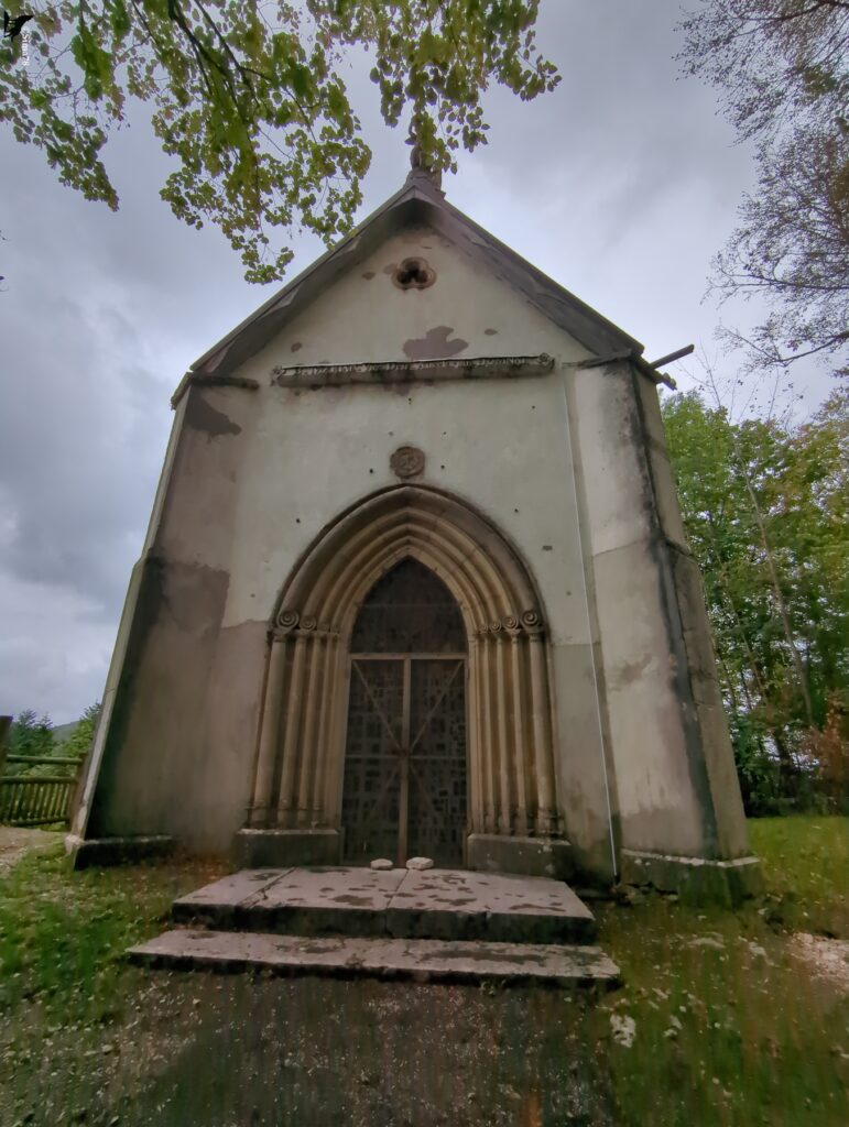 Petite chapelle au pignon crépi clair, percée d’un portail en arc brisé aux voussures sculptées et fermée par une grille, précédée de deux marches de pierre, avec un rosace trilobée au-dessus de l’entrée, entourée d’arbres et d’un ciel nuageux.